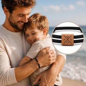 May include: A black bracelet with a wooden square charm featuring a sun, heart, and the letters A and S. The bracelet is worn on a person's wrist. The background shows a man and a child hugging on a beach.