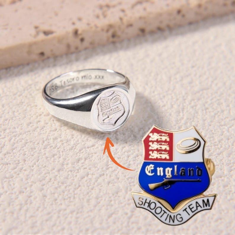 May include: A silver signet ring with an engraved shield crest and the inscription "SB Tesoro mio xxx" on the inner band, paired with a colorful enamel pin featuring the England Shooting Team crest with three lions, a target, and a rifle.