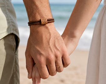 May include: A brown bracelet with a wooden square accent featuring initials and a cross design. The bracelet is worn on a wrist, with another person's hand holding it. A gold ring is visible on a finger.