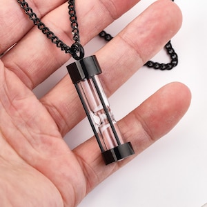 Pode incluir: Um colar de corrente preta com um pingente de ampulheta preto. A ampulheta é feita de vidro e tem uma moldura de metal preto.