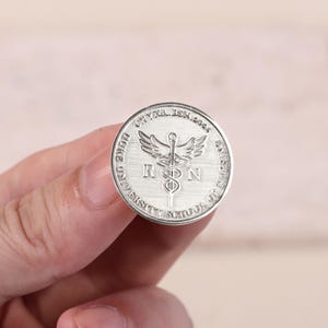 以下が含まれることがあります： 縁に「Duke University School of Nursing」と「CYNTHIA BSN 2024」の文字が刻まれた銀貨。中央には「RN」の文字が入ったカドゥケウスのシンボルがあります。