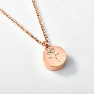 Può includere: Collana color oro rosa con un ciondolo rotondo con un disegno floreale nero. Il ciondolo è attaccato a una catena abbinata. La collana è presentata su sfondo bianco. La catena è lunga circa 50 cm.