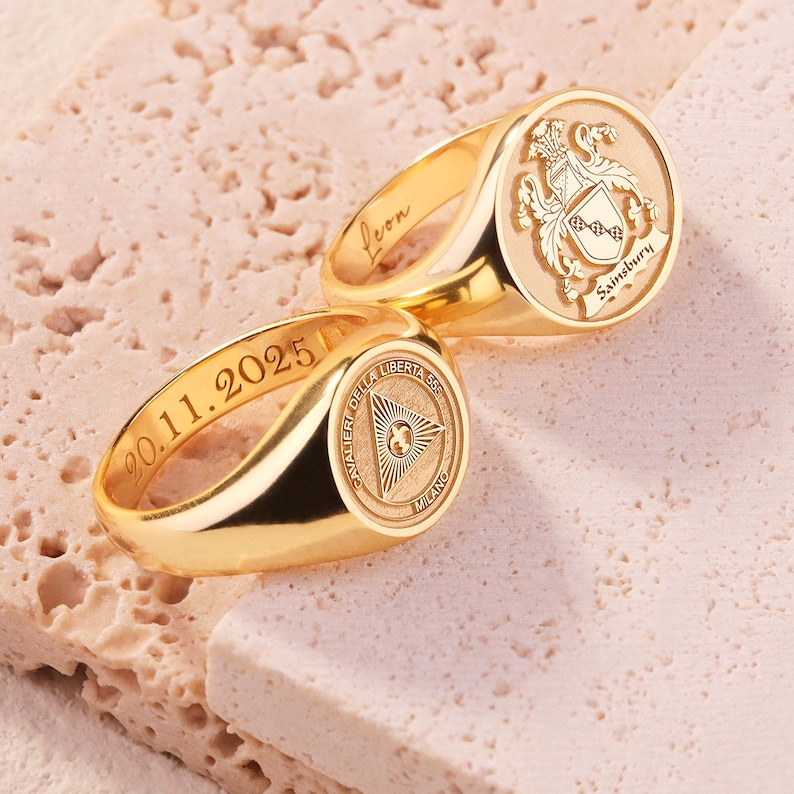 May include: Two gold signet rings with engraved details. One ring features a crest with the name Sainsbury and the interior name Leon. The other ring displays a triangular emblem with the text Cavalier della Liberta 55 Milano and the date 20.11.2025.