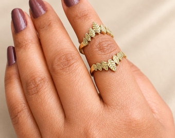 May include: Two gold-toned rings worn on the same finger, each featuring a V-shaped band adorned with a series of small, textured leaf-shaped elements arranged in a row. The rings have a matte finish and a pointed design that follows the finger.