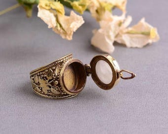 Könnte beinhalten: Ein goldener Ring mit einem Scharnierdeckel, der sich öffnet und ein kleines Fach freigibt. Der Ring ist mit filigranen Blumendesigns verziert.