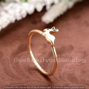 Può includere: Un anello dorato con un piccolo design a forma di coniglio. Il coniglio è rivolto a destra e ha una coda leggermente sollevata.