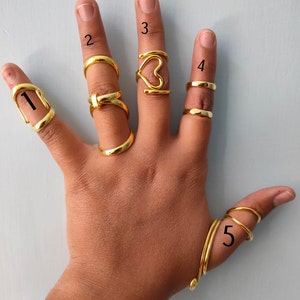 May include: A hand displaying five gold-colored rings. The rings include a full finger ring, a band ring, a heart-shaped ring, a double band ring, and a spiral ring. The rings are displayed on a light blue background.