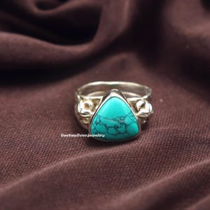 以下が含まれることがあります： 三角形のトルコ石をあしらったシルバーリング。リングには花の模様が施された装飾的なバンドがあります。トルコ石は、濃い色の縞模様が入った鮮やかな青緑色です。リングは茶色の背景に置かれています。