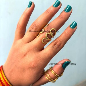 May include: Gold-toned rings in various designs adorn a hand with teal nail polish. One ring features a heart-shaped design, while another wraps around the thumb with multiple bands. The text "Geetasilverjewelry" is visible on the image. Jewelry.