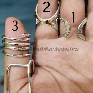 Könnte beinhalten: Drei silberne Ringe an einer Hand. Der Ring am Zeigefinger ist ein schlichter Ring mit einem geschwungenen Design. Der Ring am Mittelfinger ist ein dicker Ring mit einem geometrischen Design. Der Ring am Daumen ist ein Stapel aus vier dünnen Ringen.