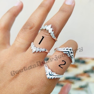 Könnte beinhalten: Silberringe mit dekorativen Designs werden auf Fingern gezeigt. Ring 1 hat ein spitzes, blattartiges Design. Ring 2 hat ein abgerundetes, perlenbesetztes Design. Die Ringe befinden sich auf hellem Hintergrund.