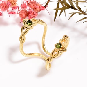 Peut inclure: Une bague en or avec un motif de nœud celtique et deux pierres précieuses vertes. La bague est ouverte et peut être ajustée pour s'adapter à différentes tailles de doigts.