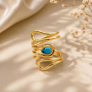 May include: A gold-toned ring featuring a continuous, fluid, and wavy metal band design. The ring is accented with a single, small, teardrop-shaped turquoise stone set within the center of the looping, polished gold-colored metal structure.