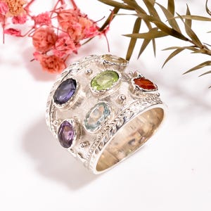 Könnte beinhalten: Ein Silberring mit einem breiten Band, das mit einem strukturierten Design und mehreren ovalen Edelsteinen in verschiedenen Farben verziert ist, darunter Amethyst, Aquamarin, Peridot, Citrin und Karneol.
