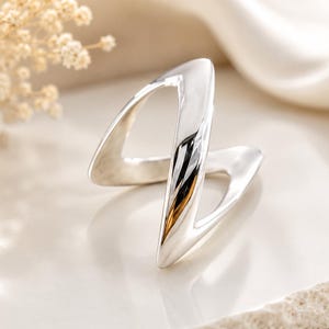 May include: A polished silver ring featuring a continuous, open-work design with two intersecting V-shaped segments that create a dynamic, angular silhouette. The metal surface is smooth and reflective, showcasing a bright, metallic finish throughout.