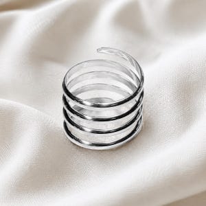 Op de afbeelding: Een zilverkleurige ring met een spiraalvormig ontwerp. De ring is gemaakt van meerdere banden die om elkaar heen wikkelen, waardoor een gelaagd effect ontstaat. De ring staat tegen een witte, getextureerde achtergrond.