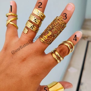 May include: Five gold-toned rings, each with a different design. The rings are worn on a person's fingers. The rings are stacked on top of each other.