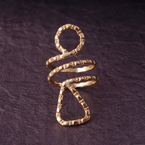 May include: A gold-colored, spiral ring with a unique design. The ring features a circular top, a triangular bottom, and a spiral band. The ring is made of a textured metal and is set against a dark background.