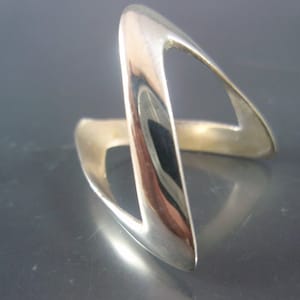 Puede incluir: Un anillo de tono plateado pulido con un diseño abierto y angular, con una forma continua y fluida que crea una silueta en zigzag. La superficie metálica es lisa y reflectante, lo que le otorga un aspecto brillante y metálico.