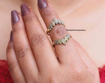 May include: Two gold-toned rings worn on a finger, each featuring a chevron-shaped band adorned with a series of small, textured leaf-shaped metal elements. The rings have a metallic finish and are positioned one above the other on the finger.