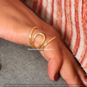 Puede incluir: Un anillo dorado con un diseño en espiral único. El anillo se envuelve alrededor del dedo con una superficie texturizada. La joya se muestra en una mano con manicura roja. El texto "Geetasilver jewelry" es visible.