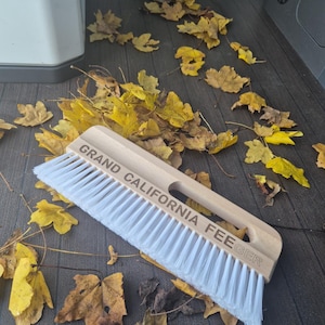 Könnte beinhalten: Eine Holzbürste mit weißen Borsten und dem Text "GRAND CALIFORNIA FEEGER" ist von gefallenen gelben und braunen Blättern umgeben. Die Bürste liegt auf einer dunkelgrauen Oberfläche.