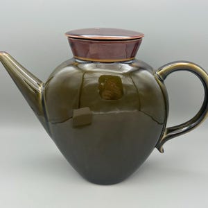 May include: A ceramic teapot with a dark olive green body and a dark brown lid. The teapot has a curved handle and a spout. The teapot is a decorative item.