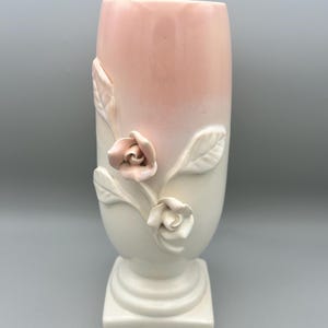 May include: A ceramic vase with a square base and a cylindrical body. The top half is a soft pink, fading into white. Three-dimensional roses and leaves in a matching pink and white adorn the front, creating a floral design. The vase is approximately 25 cm tall.