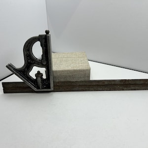 Puede incluir: Una escuadra combinada vintage, una herramienta utilizada en carpintería y metalurgia. La escuadra está hecha de metal oscuro y presenta una regla y una cabeza deslizante con un nivel y un trazador. La herramienta está diseñada para medir y marcar ángulos.