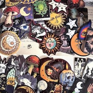 Op de afbeelding: Een verzameling diverse stickers met hemelse, mystieke en natuurlijke illustraties. Ontwerpen bevatten zonnen, manen, sterren, paddenstoelen, schedels, katten en tarot-achtige afbeeldingen in verschillende kleuren, vormen en maten op papier.