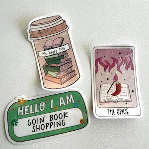 May include: Three stickers with book-themed designs. One sticker is a pill bottle labeled "My Happy Pills" filled with books. Another says "Hello I Am Goin' Book Shopping". The third is a tarot card with a chili pepper and the text "The Spice".