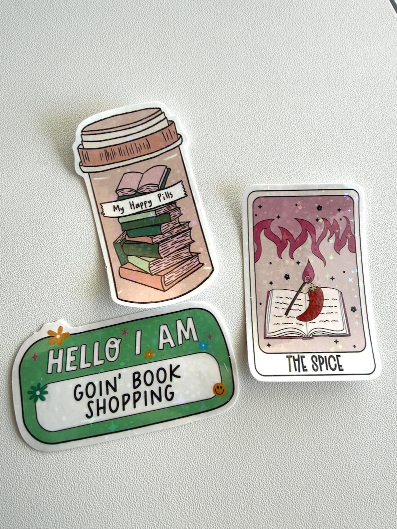 May include: Three stickers with book-themed designs. One sticker is a pill bottle labeled "My Happy Pills" filled with books. Another says "Hello I Am Goin' Book Shopping". The third is a tarot card with a chili pepper and the text "The Spice".