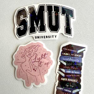May include: A collection of stickers with various designs. One sticker reads "SMUT UNIVERSITY" in black and white. Another features a pink illustration with the text "Light it up." The third sticker is a stack of books with titles like "Dark Romance."