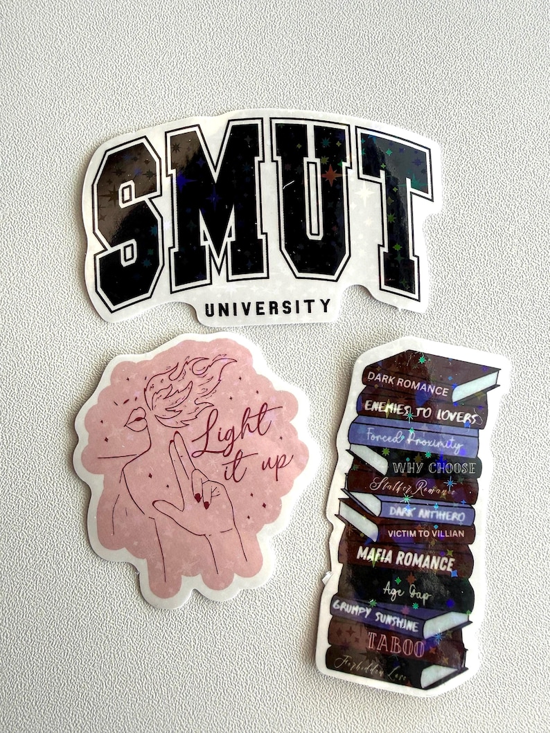 May include: A collection of stickers with various designs. One sticker reads "SMUT UNIVERSITY" in black and white. Another features a pink illustration with the text "Light it up." The third sticker is a stack of books with titles like "Dark Romance."