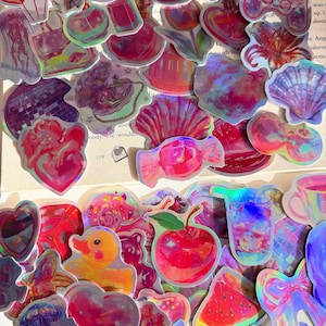 Könnte beinhalten: Eine Sammlung holografischer Aufkleber mit verschiedenen Motiven wie Herzen, Blumen, Muscheln, einer Quietscheente, einer Kirsche, einem Gamecontroller, einer Discokugel und einem Stück Wassermelone, die alle einen lebendigen, irisierenden Farbeffekt zeigen.