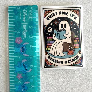 May include: A rectangular sticker featuring a cartoon ghost reading a book in a chair, with the text "QUIET NOW, IT'S READING O'CLOCK". The sticker is approximately 3 inches tall and 2 inches wide.