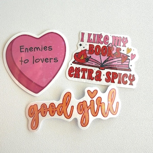 May include: Three stickers with text on a white background. One is a pink heart with "Enemies to lovers" in black. Another says "I like my books extra spicy" with a book and chili pepper graphic. The third says "good girl" in orange.