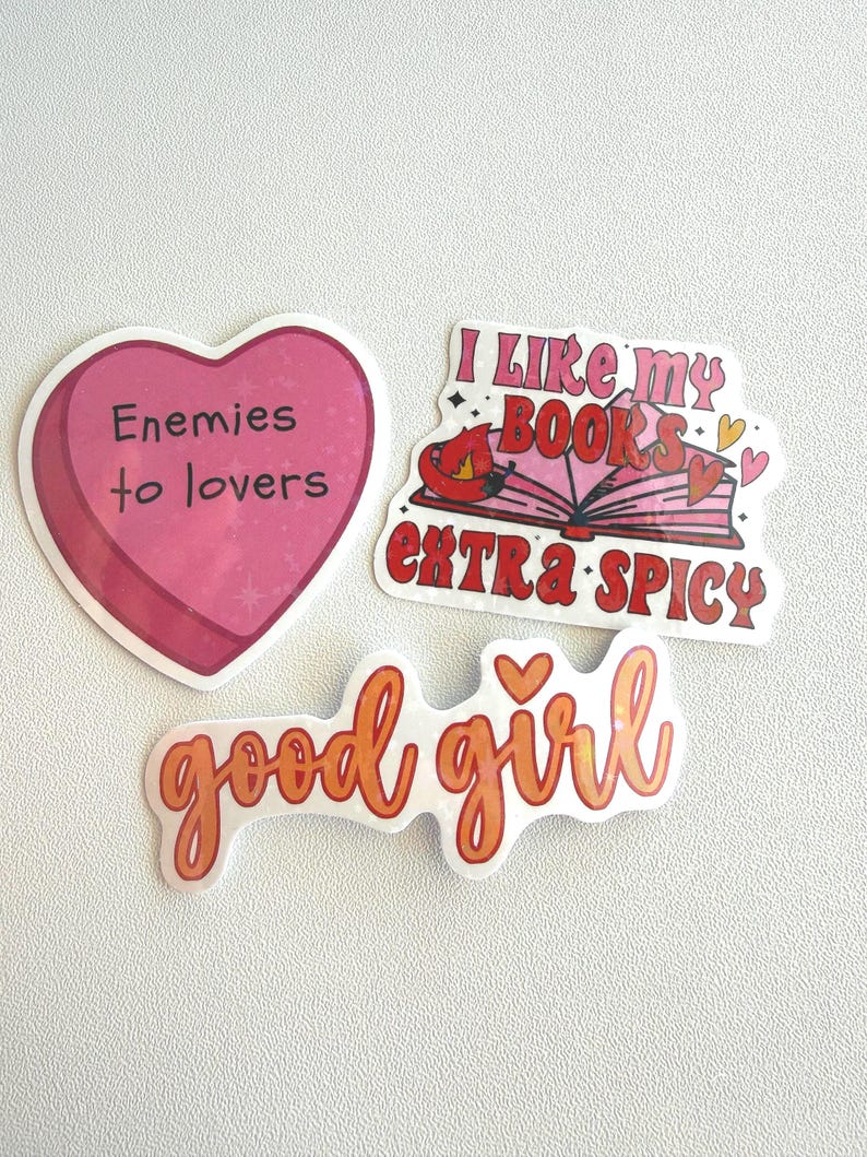 May include: Three stickers with text on a white background. One is a pink heart with "Enemies to lovers" in black. Another says "I like my books extra spicy" with a book and chili pepper graphic. The third says "good girl" in orange.
