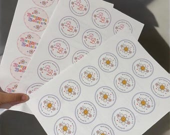 May include: Sheets of round stickers with various designs. One sheet features the text "Business Basics &" in pastel colors. Other sheets display a "BB" monogram and a smiling daisy design, all within circular frames. The stickers are for a creative studio.