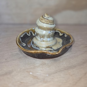 May include: A ceramic ring holder featuring a circular, dark brown glazed dish with a wavy rim and a central, spiraled, cream-coloured cone. The glaze on the dish shows lighter brown drips, while the cone has a textured, layered appearance.