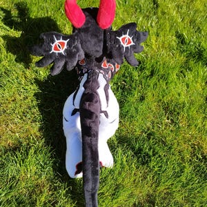 May include: A plush toy in the shape of a fantasy creature, featuring red horns, black wings with eye designs, and a long black tail. The toy is primarily black and white, with red accents, and is set against a green grassy background.