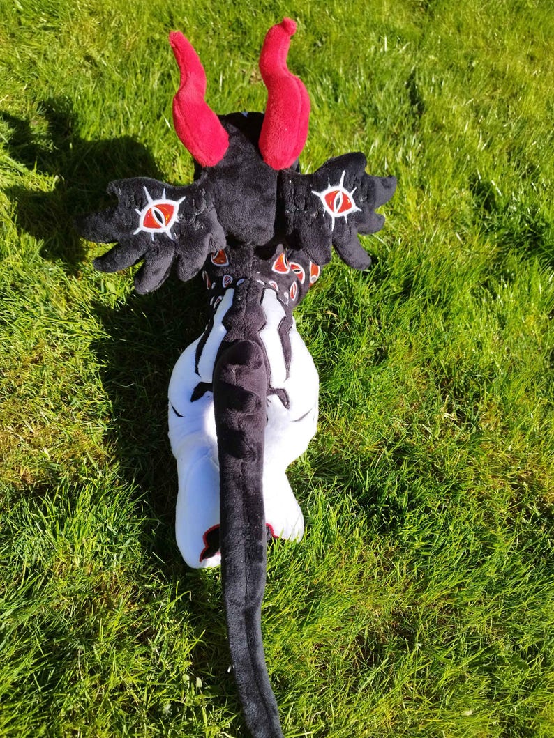 May include: A plush toy in the shape of a fantasy creature, featuring red horns, black wings with eye designs, and a long black tail. The toy is primarily black and white, with red accents, and is set against a green grassy background.