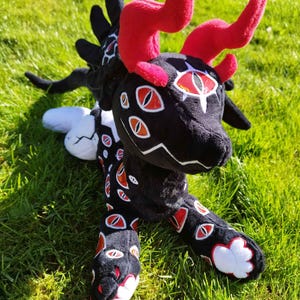 May include: A plush creature with a black body, red horns, and multiple red and white eye designs. The plush has a long tail and is lying on green grass. The creature's horns are a vibrant red, contrasting with the black body.