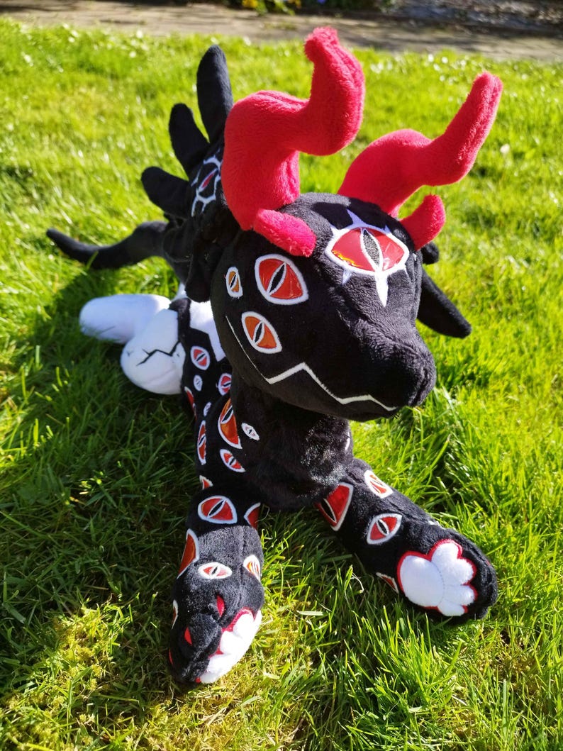 May include: A plush creature with a black body, red horns, and multiple red and white eye designs. The plush has a long tail and is lying on green grass. The creature's horns are a vibrant red, contrasting with the black body.