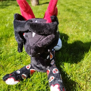 May include: A plush toy dragon with a black body, red horns, and white accents. The dragon's legs are decorated with red and white eye-shaped patterns. The toy is positioned on a green grassy surface.