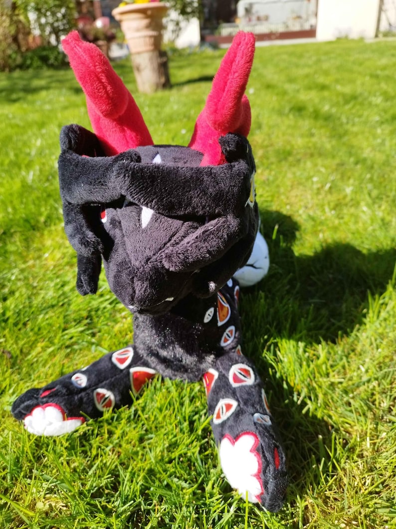 May include: A plush toy dragon with a black body, red horns, and white accents. The dragon's legs are decorated with red and white eye-shaped patterns. The toy is positioned on a green grassy surface.