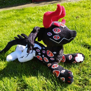 May include: A black plush creature with red accents, including horns and eye-shaped designs. The plush toy has multiple red and white eyes and is lying on green grass. The creature's paws have white accents.