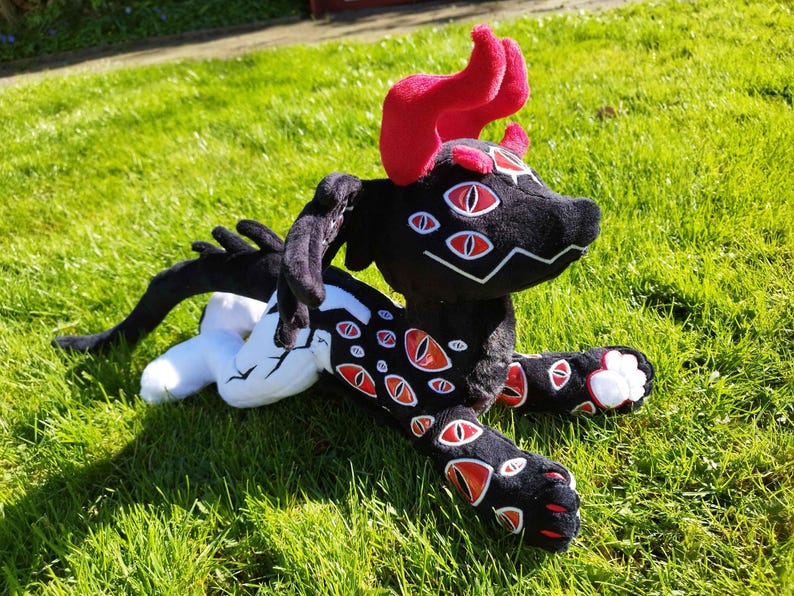 May include: A black plush creature with red accents, including horns and eye-shaped designs. The plush toy has multiple red and white eyes and is lying on green grass. The creature's paws have white accents.