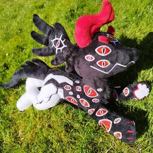 May include: A plush toy dragon with a black body, white accents, and multiple red-and-white eyes. The dragon has red horns and is lying on green grass. The toy is soft and detailed, with a whimsical design.