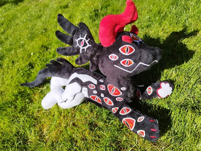 May include: A plush toy dragon with a black body, white accents, and multiple red-and-white eyes. The dragon has red horns and is lying on green grass. The toy is soft and detailed, with a whimsical design.